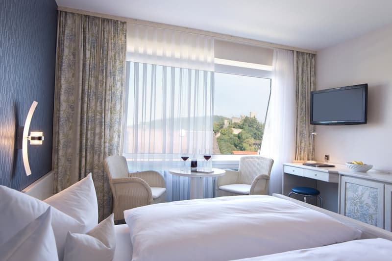 Spacious bedroom with large windows offering a scenic view, a round table with two chairs, a TV mounted on the wall, and a dresser with drawers.