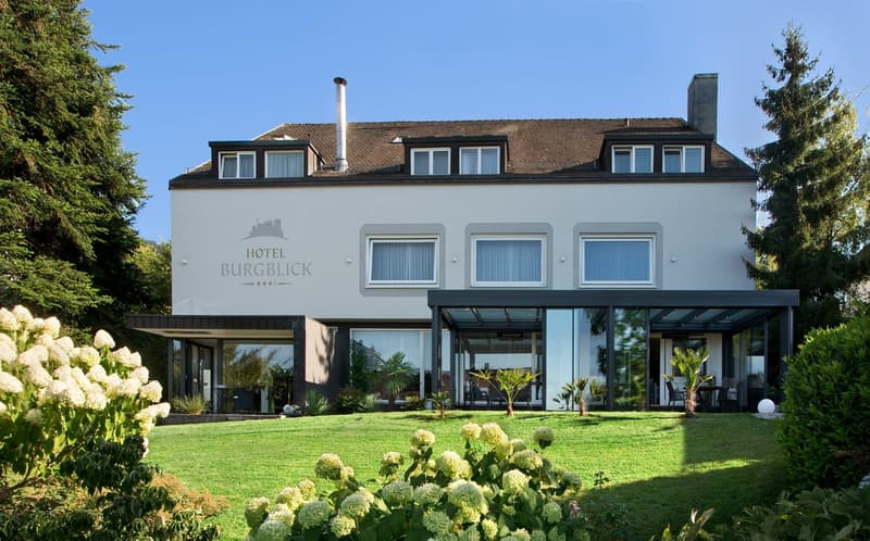 4-star Hotel Burgblick, 3-story building with a brown tiled roof, large windows, and a glass-enclosed patio area. The hotel is surrounded by a well-manicured lawn and lush landscaping, including flowering plants and shrubs.
