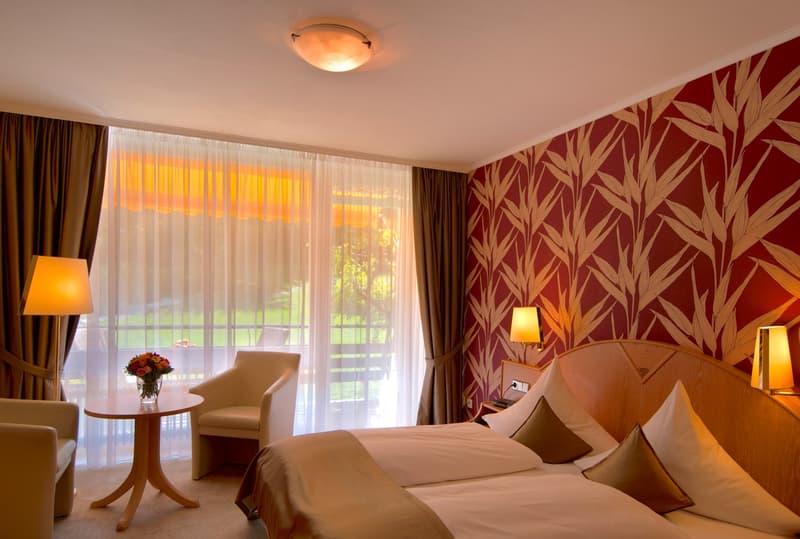 Spacious bedroom with a large window overlooking a balcony or terrace, burgundy and beige patterned wallpaper, wooden furniture including a round table and armchair, and warm lighting from a ceiling fixture and table lamps.