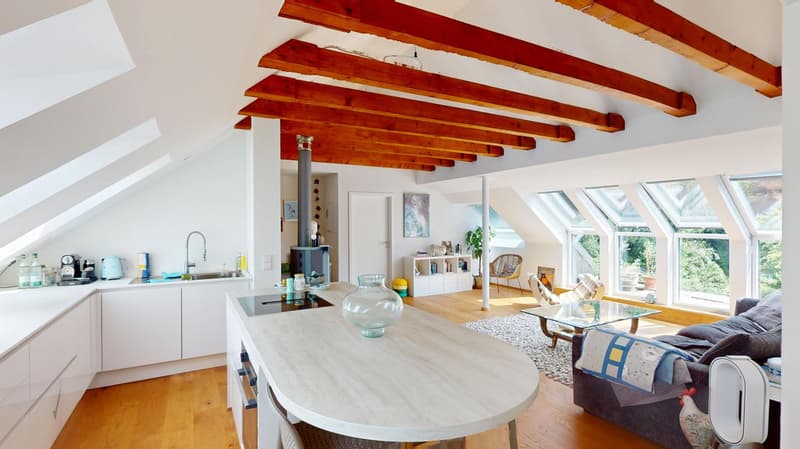 Spacious open-plan living area with a kitchen, dining table, and living room. The room features large windows providing a scenic view, and a wooden beamed ceiling. The kitchen has modern appliances and cabinets. The living room has a sofa and a glass coff