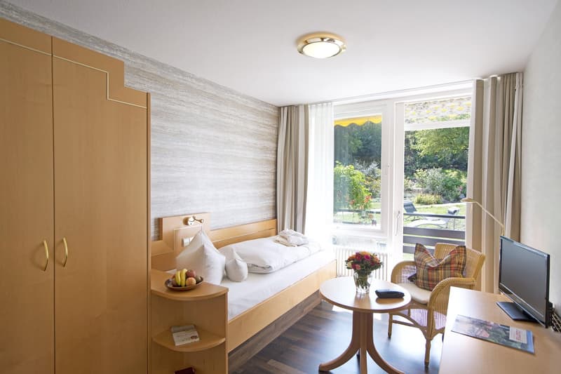 Spacious bedroom with large windows overlooking a garden, wooden furniture including a bed, nightstand, and armchair, and a balcony or terrace providing a scenic view.