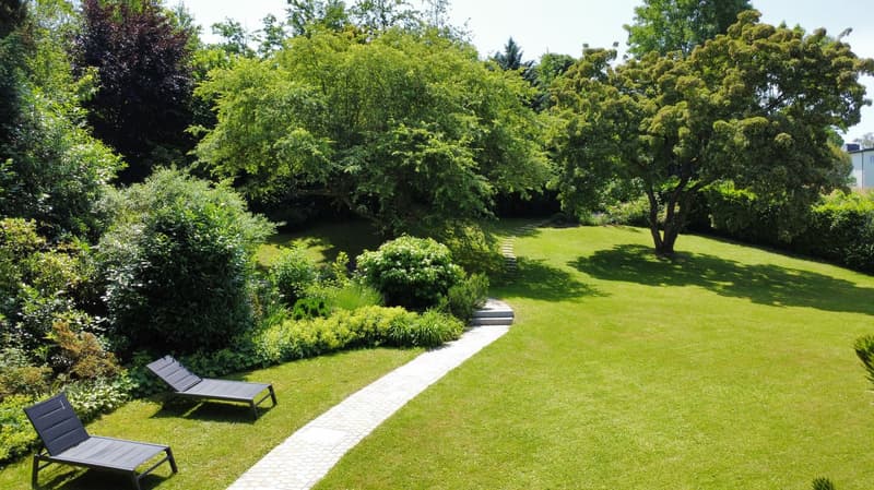 Lush green garden with a well-maintained lawn, a paved walkway, and several trees and shrubs. There are two lounge chairs visible, suggesting a relaxing outdoor space.
