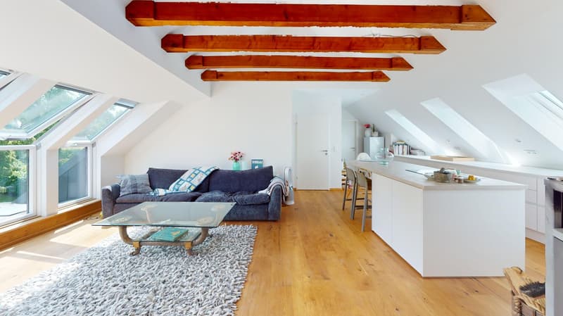 Spacious attic living room with exposed wooden beams, large windows providing natural light, grey sofa, glass coffee table, and a kitchen area with a counter and stools