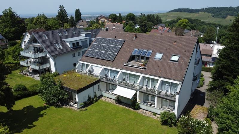 Multi-story apartment building with solar panels on the roof, balconies, and surrounded by greenery with a view of the landscape in the background.