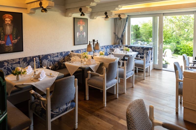 Spacious dining room with large windows overlooking a garden, wooden floors, floral patterned upholstered chairs, white tablecloths, and a framed artwork on the wall