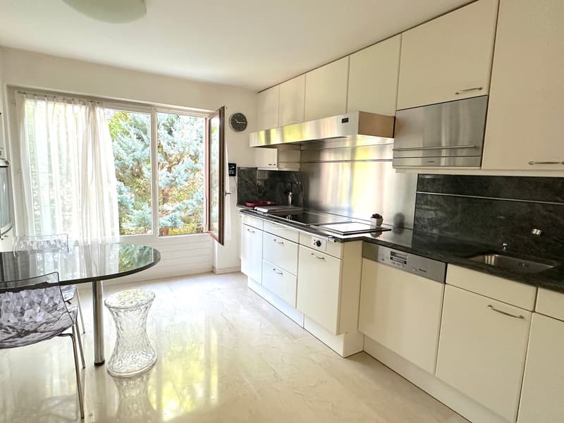 White kitchen with granite countertops, stainless steel appliances, glass table, white cabinets, large window, clock, and modern lighting