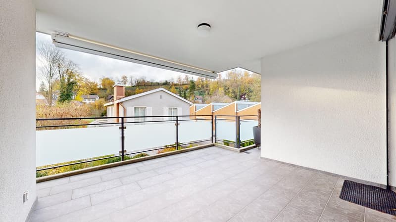 A room with tiled floor, a balcony with glass railings, overlooking the outside with a distant view of buildings and trees.