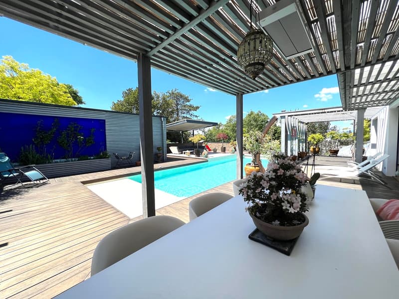 Wooden deck with dining table, covered area with pergola, swimming pool, loungers, plants, and garden wall.