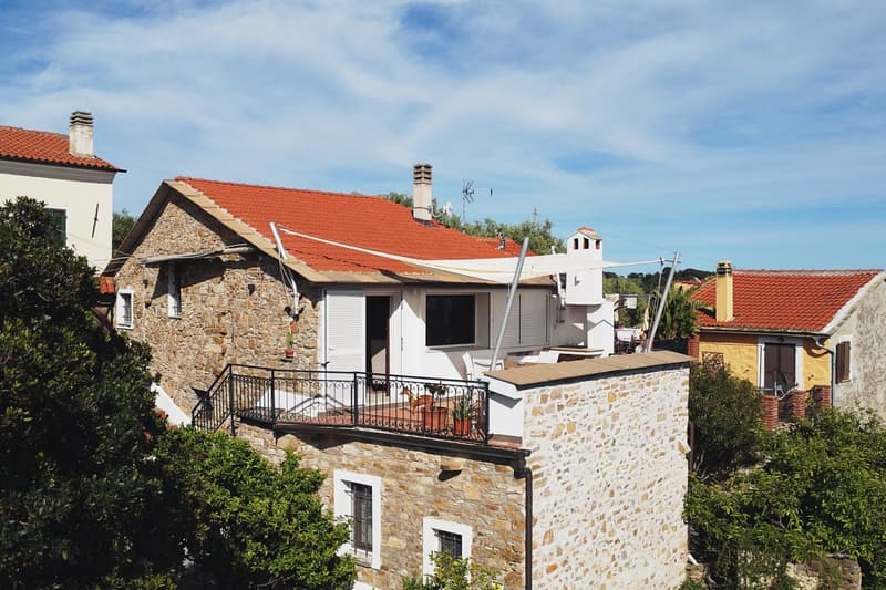 stone walls, red tiled roof, black railings on balcony, two floors, outdoor dining table, potted plants
