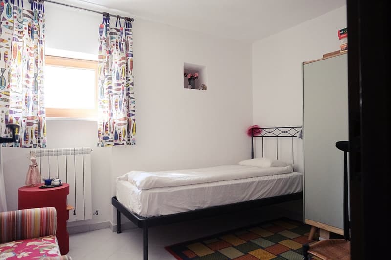 Single bed, floral pillows, wall shelf with flowers, colorful curtains, red side table, chair, radiator