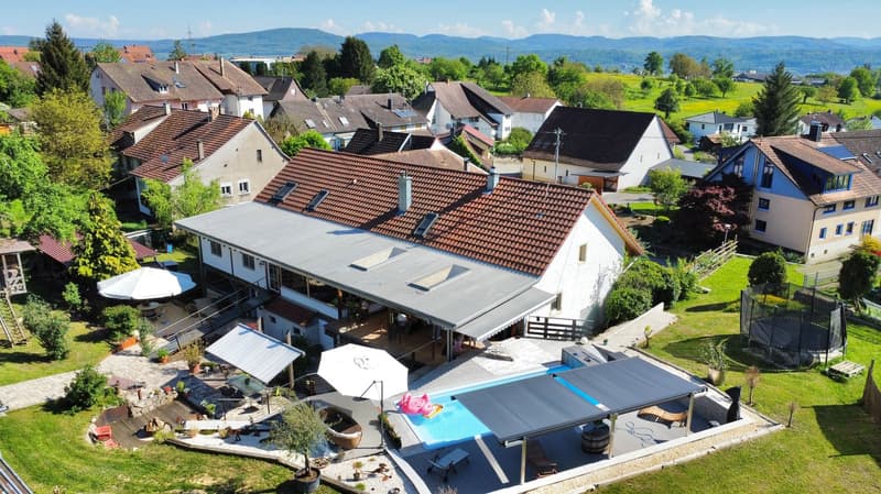Two-story house with brown roof, solar panels, outdoor swimming pool, patio with patio umbrellas, garden, nearby trampoline, and distant views of mountains.