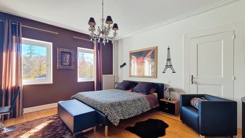 Large bedroom with bed, two night stands, armchair, chandelier, radiator, pictures on wall, wooden floor, rug
