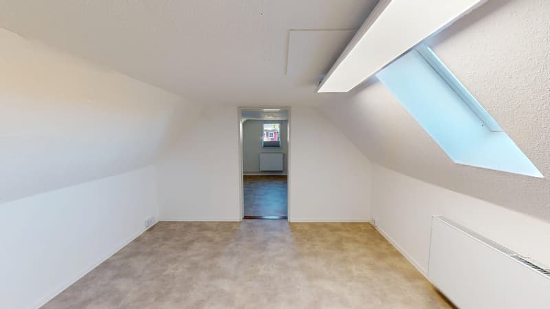 Empty room with slanted ceiling, no furniture, white walls, wooden floor, window, and heating system
