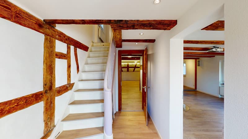White walls, wooden beams, wooden stairs, wooden floors, an open door leading to a kitchen.