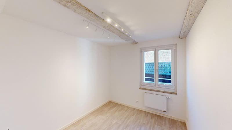Empty room with white walls and wooden flooring, window with view of a roof, ceiling light