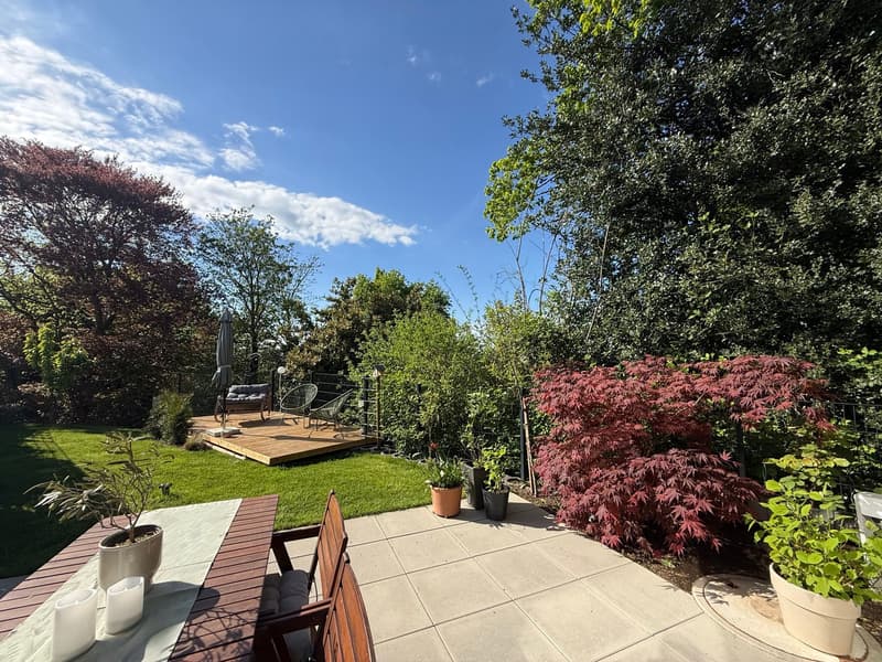 Wooden deck with chairs, plants, and a table with candles, surrounded by a green lawn and trees