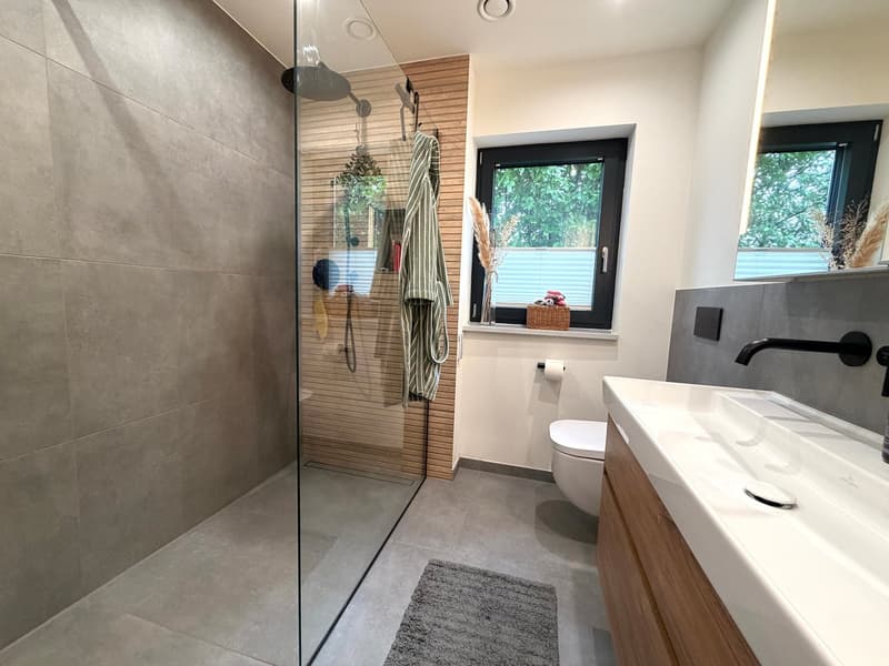 modern bathroom with glass shower, white sink, wooden cabinet, white toilet, windows with blinds, towel hanging on wall