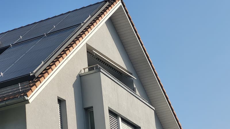 Two-story house, gray exterior, brown roof tiles, solar panels on the roof, balcony with railing, two windows, one with shutters