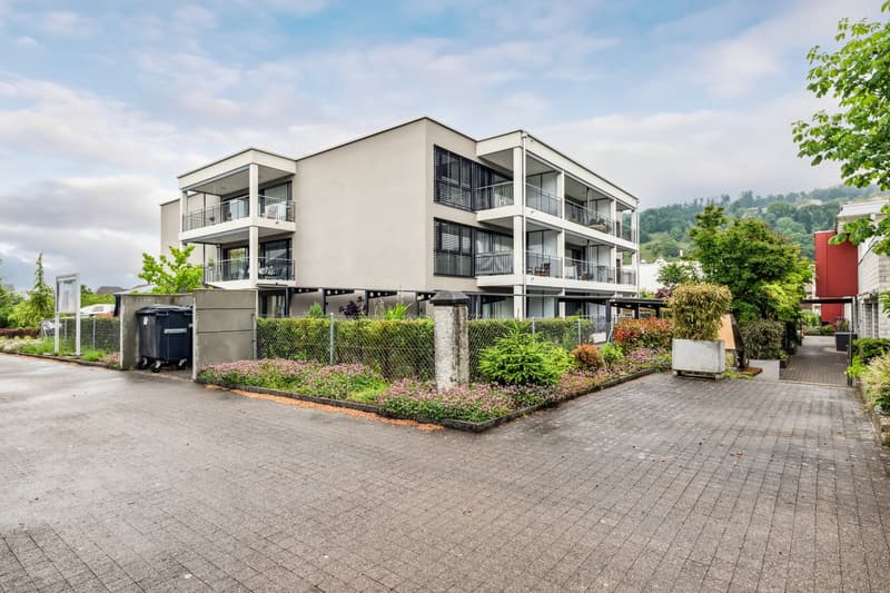 Modern apartment building with balconies, brick walls, paved driveway, greenery