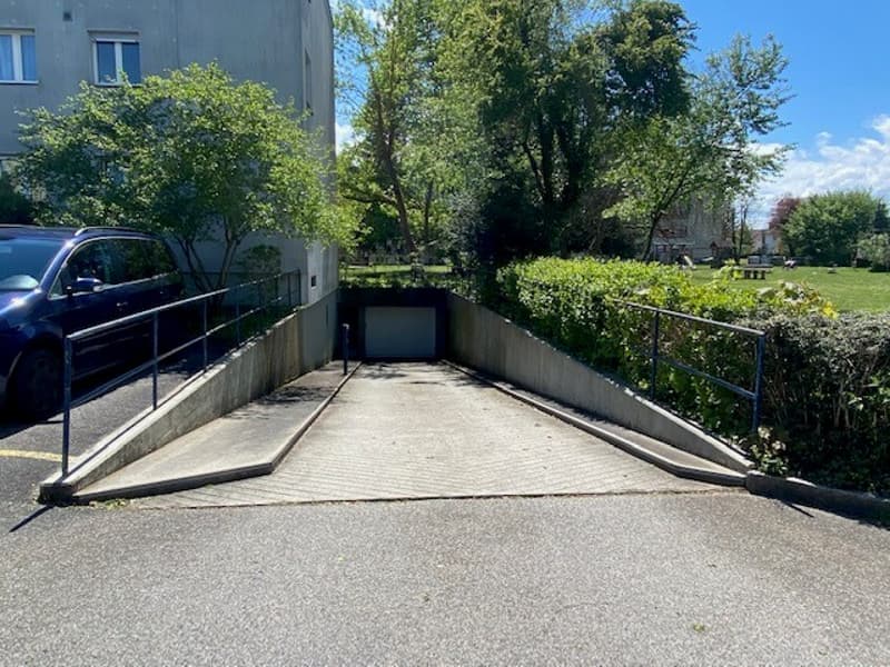 A sloping driveway leads to a garage underneath a building, with a blue car parked nearby. In the background, there is a park with greenery and a bench. The entrance is fenced with metal handrails.