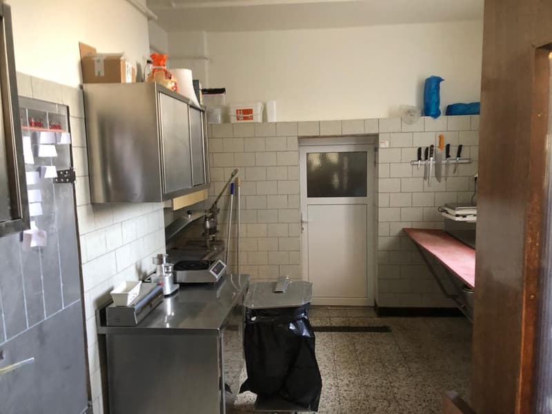 Tiled kitchen with stainless steel appliances, including a refrigerator and dishwasher. There is a counter space with various kitchen tools and equipment. The room appears to be cluttered with some boxes and other items stored in the space.
