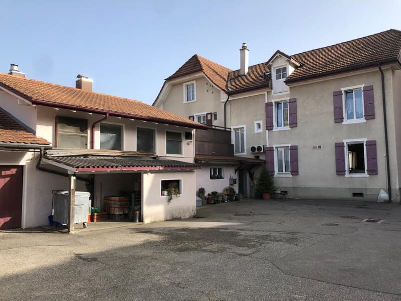 This image shows a multi-story residential building with a garage. The building has a traditional European architectural style with red tile roofs and stucco walls. There are several windows and balconies visible, and a paved courtyard or driveway in the