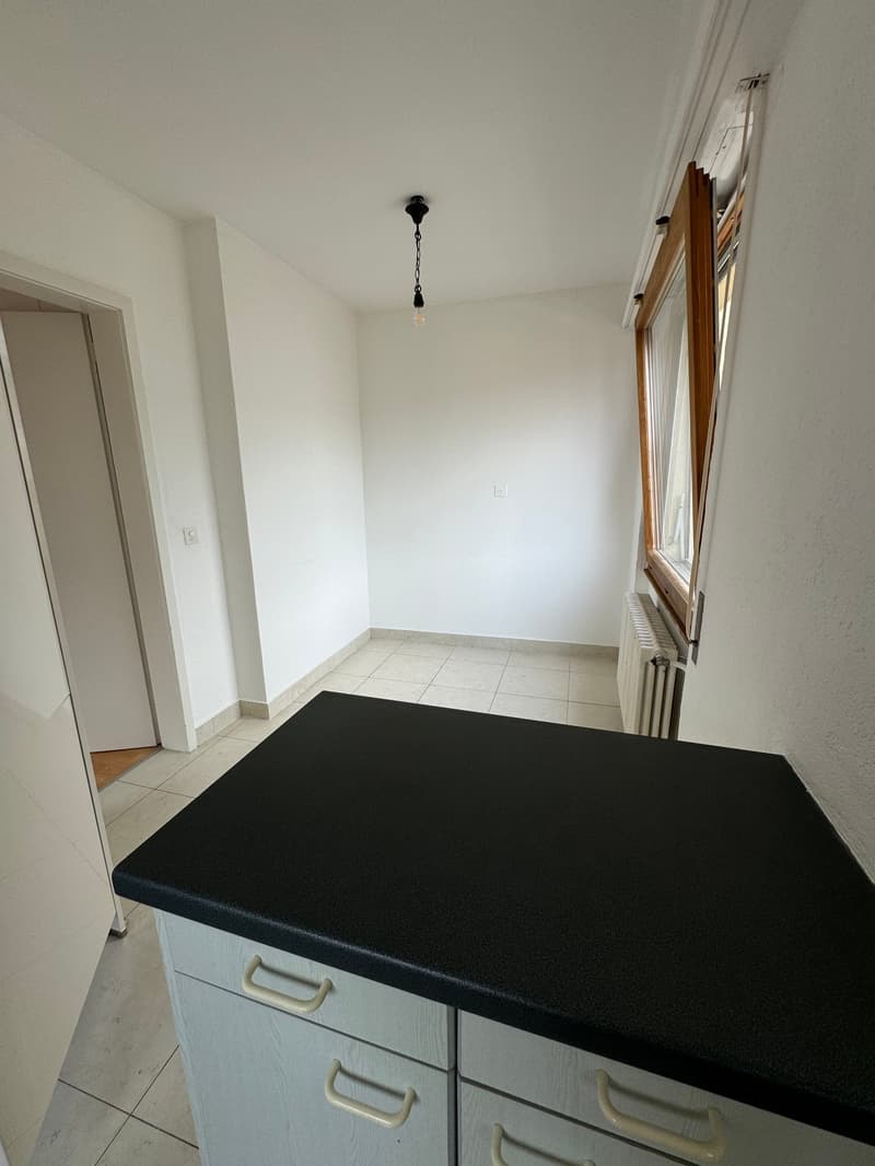 Empty room with white walls, tiled floor, and a black countertop. There are two white cabinets with handles and a window with a wooden frame.