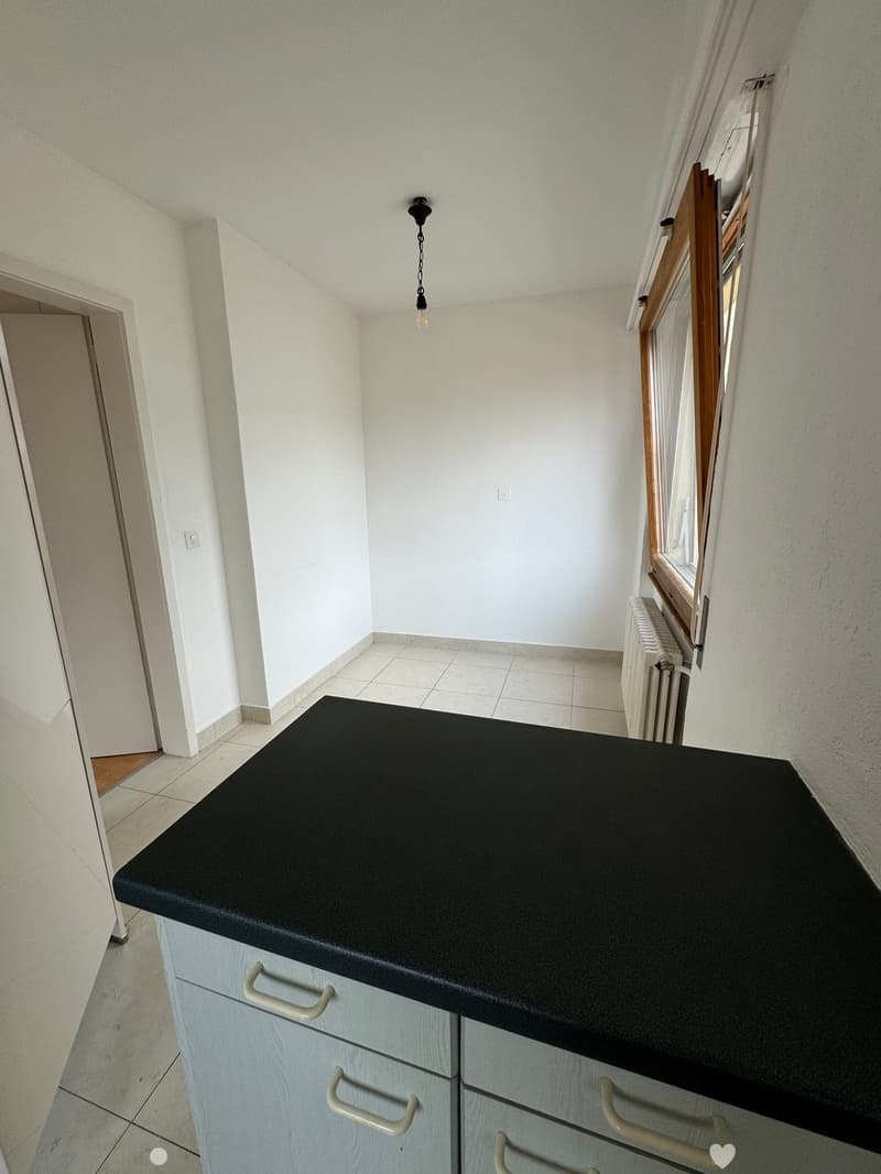 Empty room with white walls, tiled floor, and a black countertop. There are two white cabinets with handles and a window with a wooden frame.