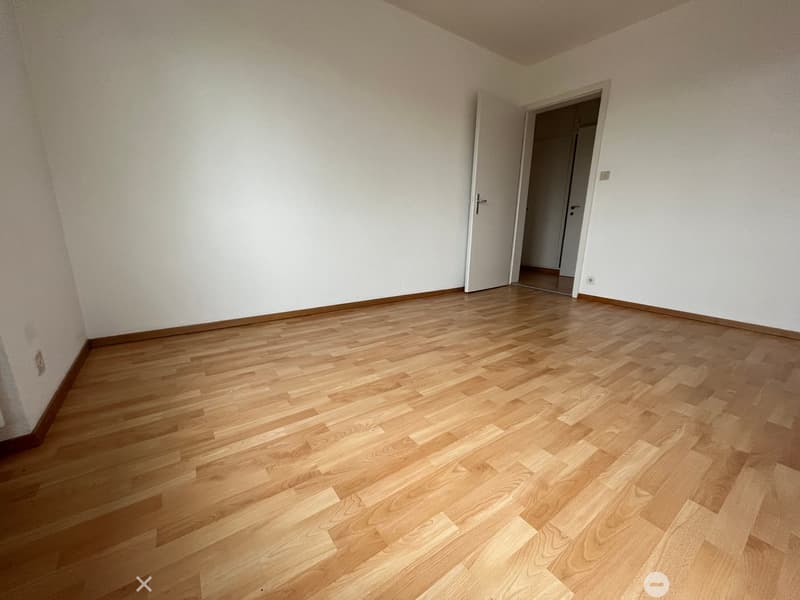 Completely empty room with white walls, wooden floor, and two doors leading to another room.