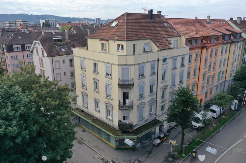 Aerial view of a multi-story building with brown roofs and multiple windows. The building is surrounded by other buildings and trees. There are vehicles parked on the side of the road.