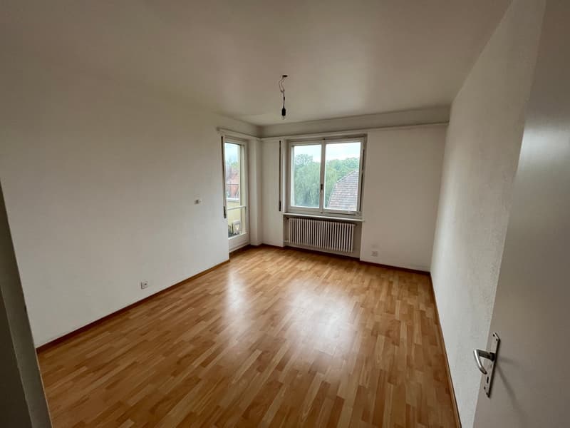 Empty room with white walls, wooden floor, and two windows with a view of trees outside.