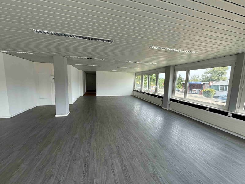 Large open commercial space with grey walls, grey wood-like flooring, and large windows providing natural light. The space appears to be a vacant office or retail unit ready for a new tenant.
