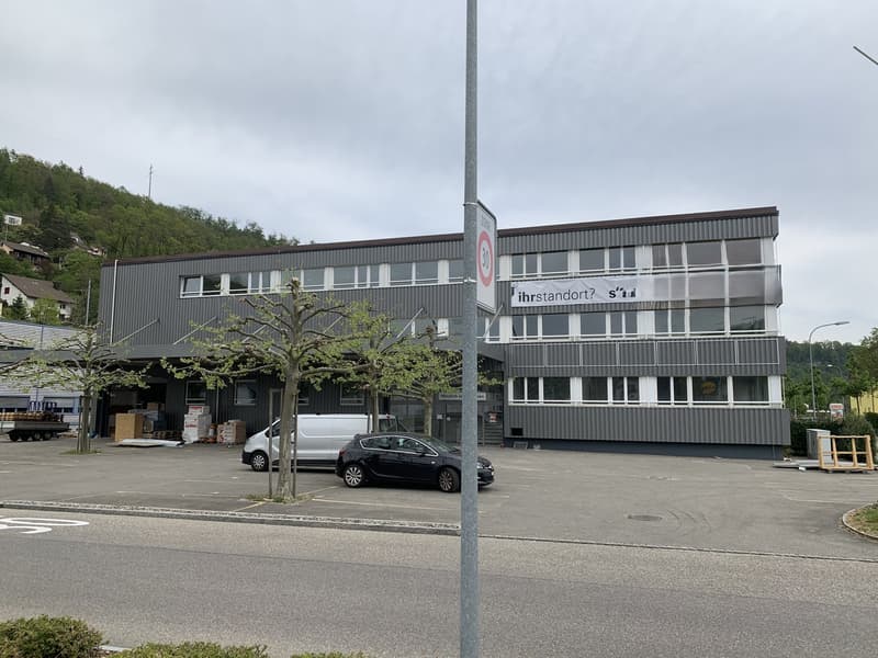 A multi-story commercial building with a large parking lot in front. The building has a grey corrugated metal exterior and several windows. There is a tree in front of the building and a few parked cars in the lot.