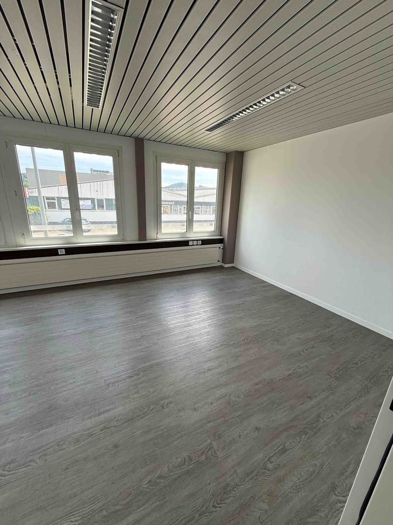 The image shows a spacious office space with a gray wood-like flooring, white walls, and a gray slatted ceiling. The room has large windows that provide natural light and a view of the surrounding buildings.