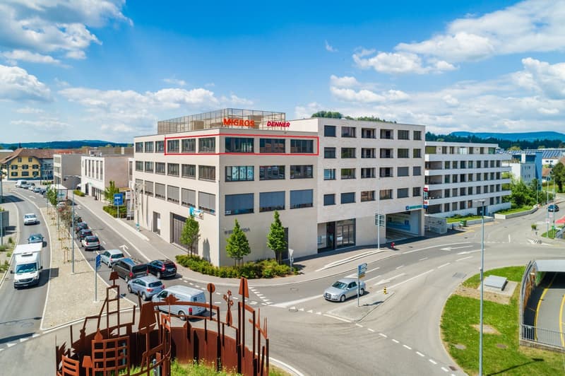 Office building, white exterior, multiple windows, multiple floors, red roof signage for Migros and Denner