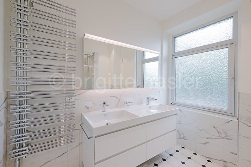 Spacious bathroom with white vanity, large mirror, and a window providing natural light. The floor features a black and white tile pattern.