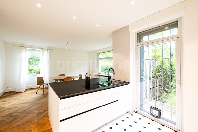 Spacious open-plan kitchen with white cabinets, black countertop, large windows overlooking a balcony or terrace, hardwood floors, and a dishwasher visible