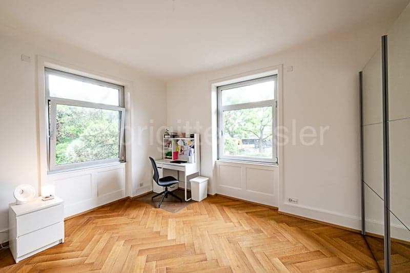 Spacious bedroom with large windows, hardwood herringbone flooring, white walls, and a desk setup near the windows