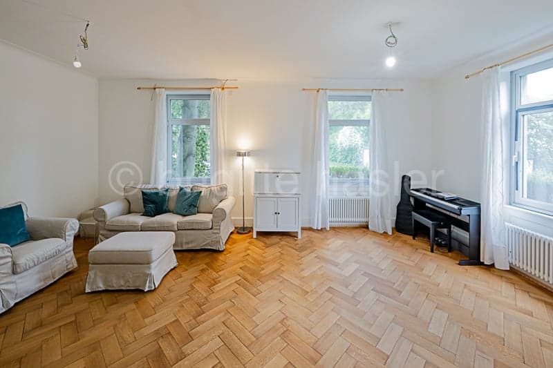 Spacious living room with hardwood herringbone flooring, large windows, white walls, beige sofa and armchair, coffee table, and a piano in the corner.