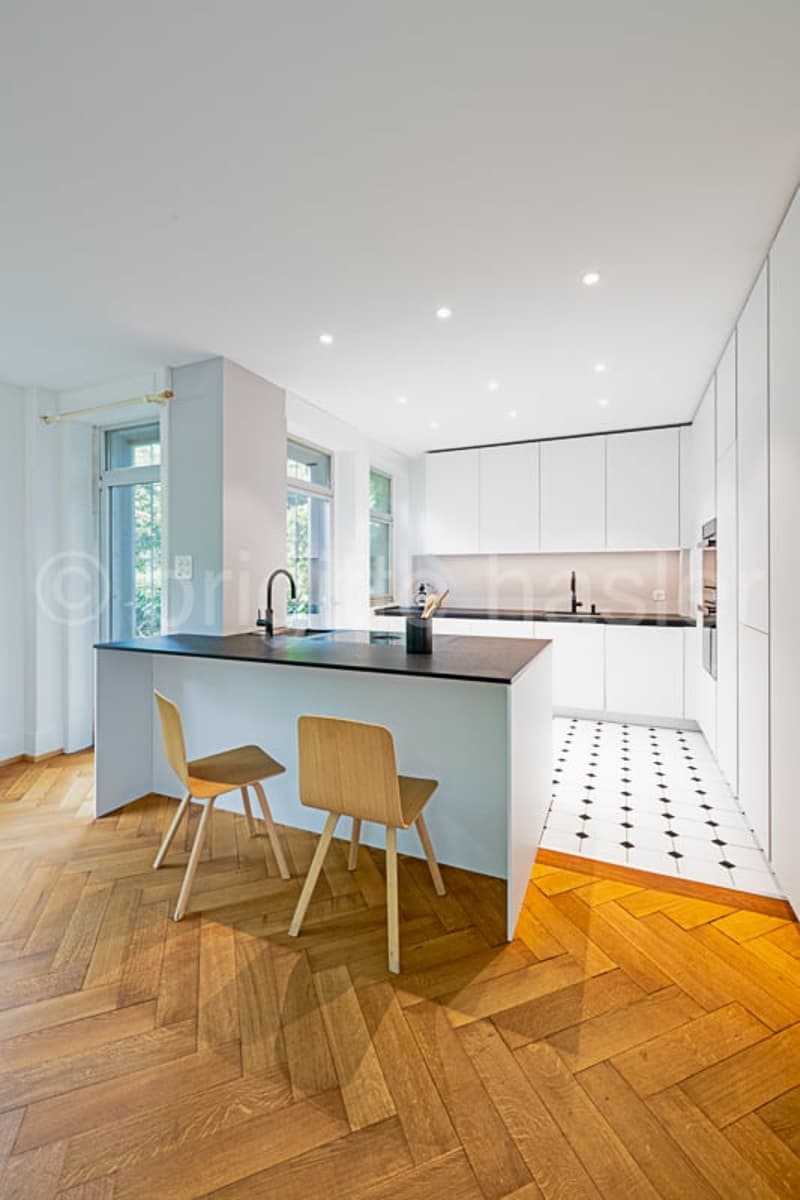Modern, open-concept kitchen with white cabinets, black countertops, and a central island with bar stools. The floor features a herringbone wood pattern. The kitchen has large windows and recessed lighting.