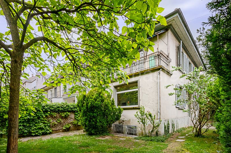 2-story house with white exterior, green trees and foliage surrounding the property, balcony on the second floor, and a well-maintained garden area in the front.