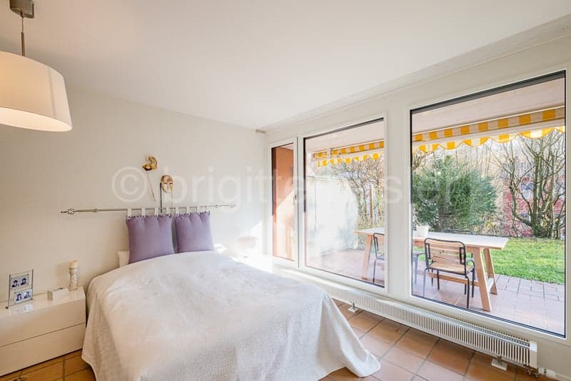 a single bed with white bed sheets and purple pillows, white nightstand with a picture frame, white walls, floor lamp, sliding glass doors, patio with wooden table and chairs, outdoor greenery and brick floor