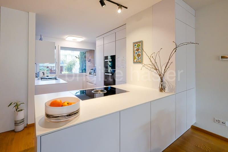 Modern kitchen with a white island, white cabinets, granite countertop, integrated oven, and gas stove. Wooden floor, decorative items on the counter, a potted plant on the floor, and an outlet on the wall.