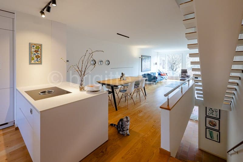 modern white kitchen, central island with cooktop, dining table with 6 chairs, hardwood floor, white walls, staircase to upper floor, living area with couch, coffee table, windows, framed pictures