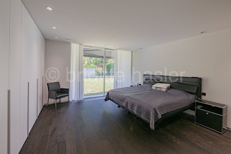 Modern design, white walls, wooden floor, bed with grey bedding, bedside tables, white curtains, black chair, large glass window overlooking a terrace with a grassy area.
