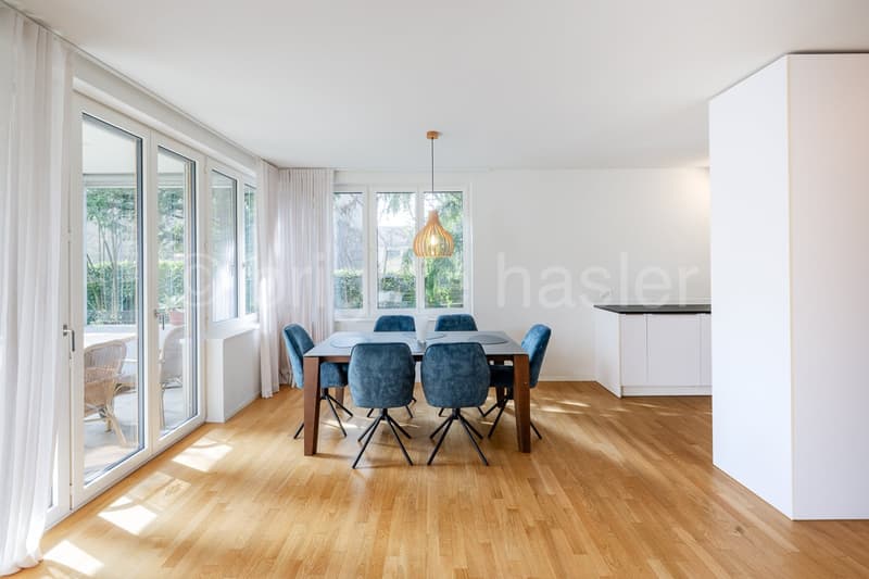 Dining room with wooden floor, large windows, glass doors, wooden table, blue chairs, pendant light, white walls, white curtains, kitchen area