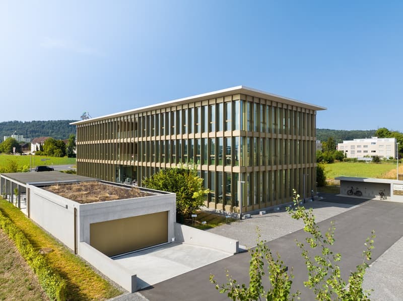 A modern, multi-story office building with a glass facade and a garage on the ground level. The building is surrounded by greenery and has a paved outdoor area with bicycle racks.