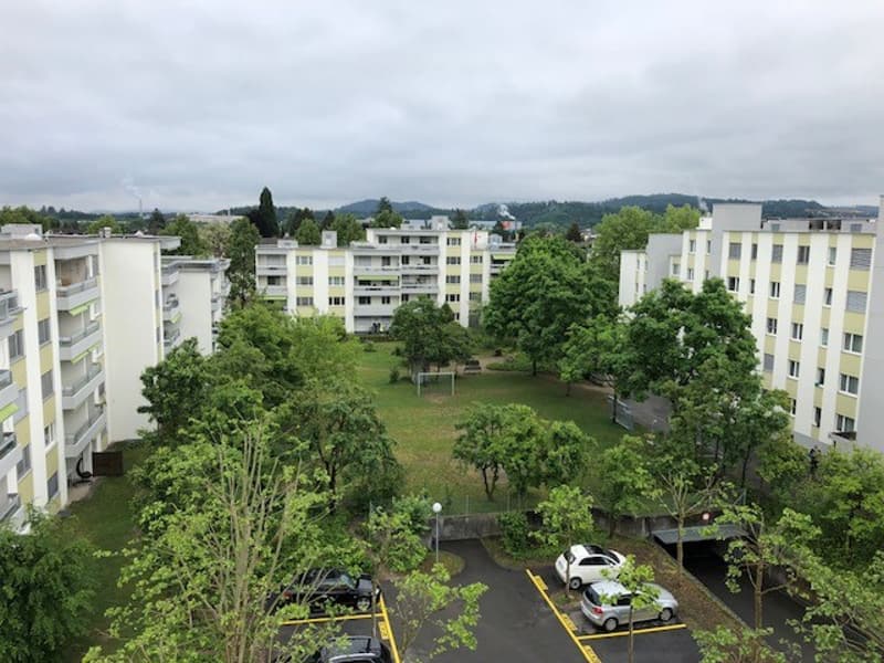 Aerial view of a residential building complex with multiple buildings, parking area, and a grassy field.