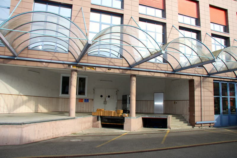 Exterior of a multi-story building with a parking area and covered entrance. A glass canopy is installed above the entrance.