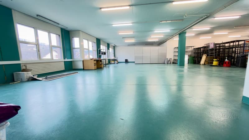 An empty room with a green floor and white walls, featuring large windows and ceiling lights.